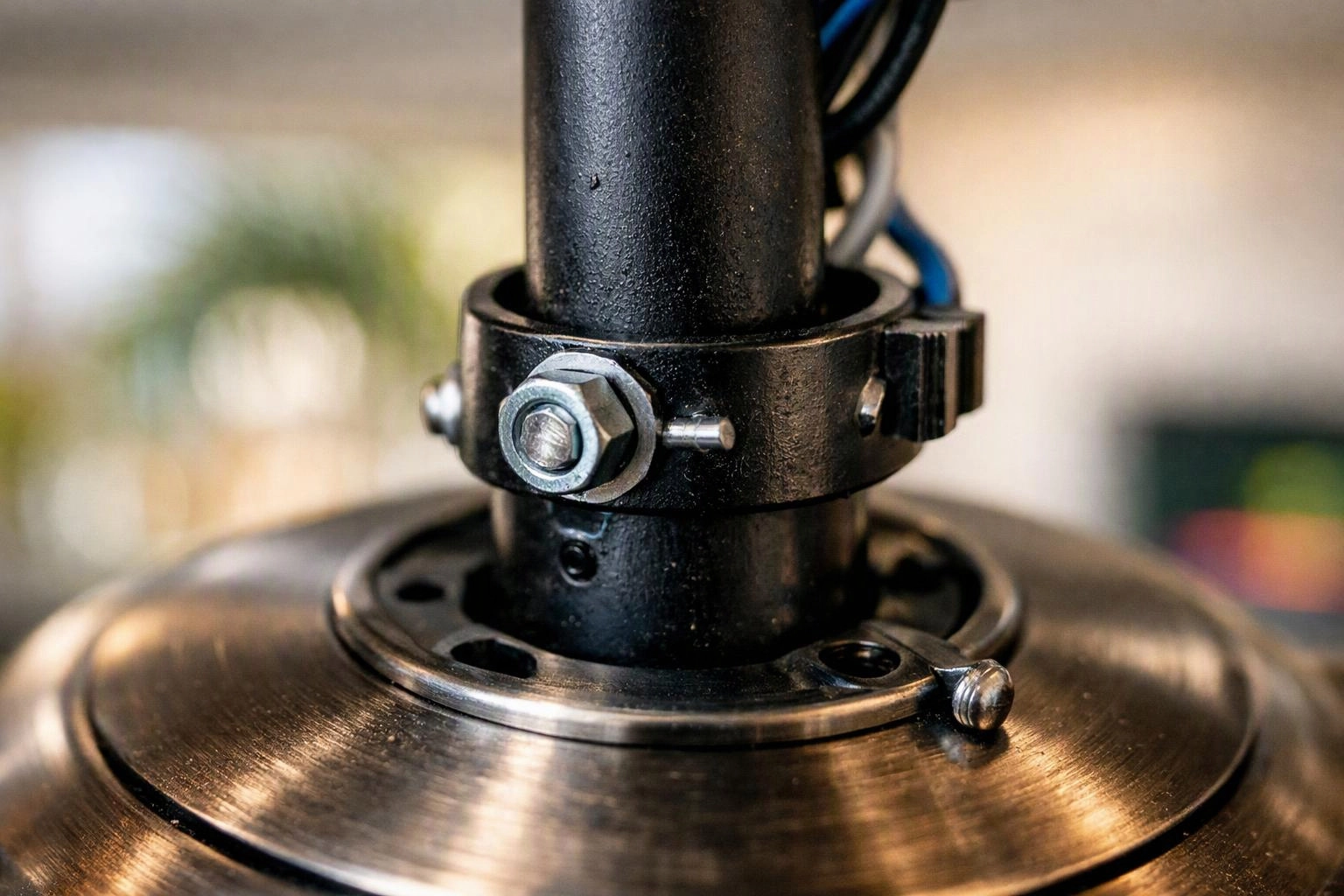 Close-up of a black metal pipe clamp and brushed metal base with bolts against a blurred indoor background